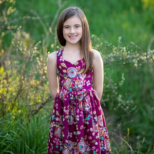 May include: A young girl wearing a sleeveless, purple dress with a floral print. The dress has a sweetheart neckline and a gathered skirt. She is standing in a field of green grass.