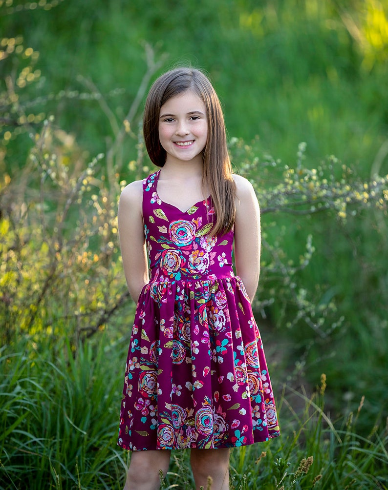 May include: A young girl wearing a sleeveless, purple dress with a floral print. The dress has a sweetheart neckline and a gathered skirt. She is standing in a field of green grass.