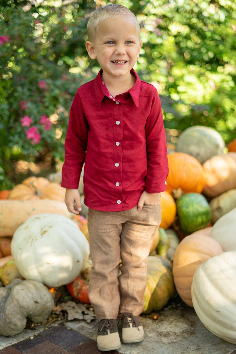 Op de afbeelding: Een jonge jongen in een rood overhemd met knopen en een bruine broek glimlacht terwijl hij voor een stapel pompoenen staat.