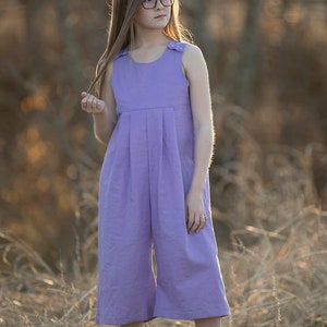 May include: A young person wearing a lavender sleeveless jumpsuit with a pleated front and button details. They are wearing glasses and standing in a field of tall grass.