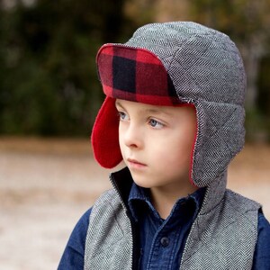 May include: A young person wearing a gray and black patterned winter hat with red and black plaid ear flaps. The hat has a brim and is pulled down over the person's forehead.