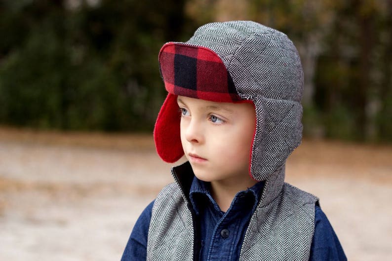 May include: A young person wearing a gray and black patterned winter hat with red and black plaid ear flaps. The hat has a brim and is pulled down over the person's forehead.