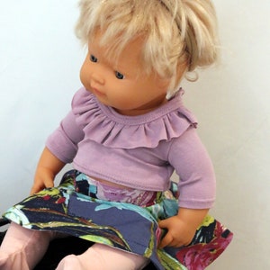 May include: A doll wearing a purple ruffled top and a floral skirt. The doll has blonde hair and is wearing a headband with a pink flower.