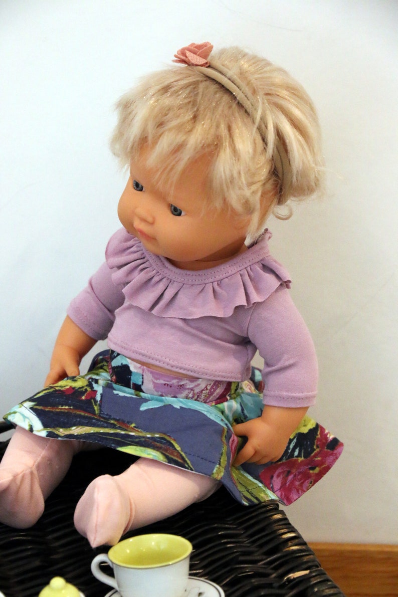 May include: A doll wearing a purple ruffled top and a floral skirt. The doll has blonde hair and is wearing a headband with a pink flower.