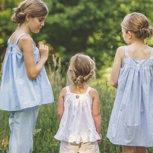 Pu&ograve; includere: Tre giovani ragazze indossano abiti senza maniche di colore azzurro chiaro con bottoni bianchi. Le ragazze sono in piedi in un campo erboso.