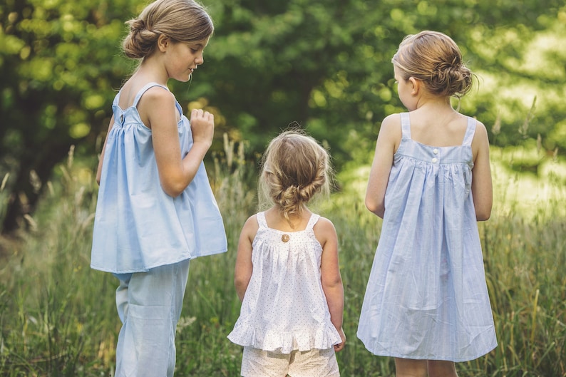 Pu&ograve; includere: Tre giovani ragazze indossano abiti senza maniche di colore azzurro chiaro con bottoni bianchi. Le ragazze sono in piedi in un campo erboso.