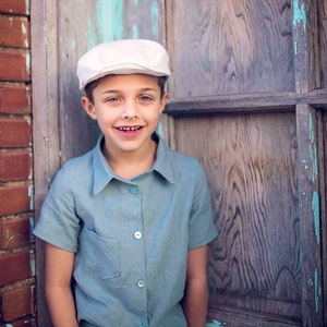 May include: A young boy wearing a white cap and a light blue button-down shirt with short sleeves. He is standing in front of a wooden door with peeling blue paint.