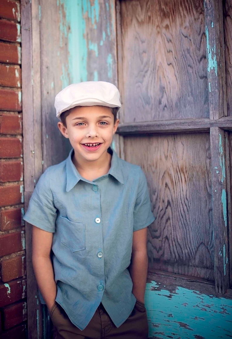 May include: A young boy wearing a white cap and a light blue button-down shirt with short sleeves. He is standing in front of a wooden door with peeling blue paint.