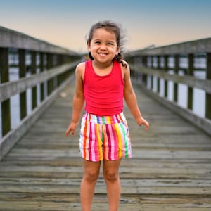 May include: A young girl wearing a red tank top and rainbow striped shorts smiles while standing on a wooden pier.