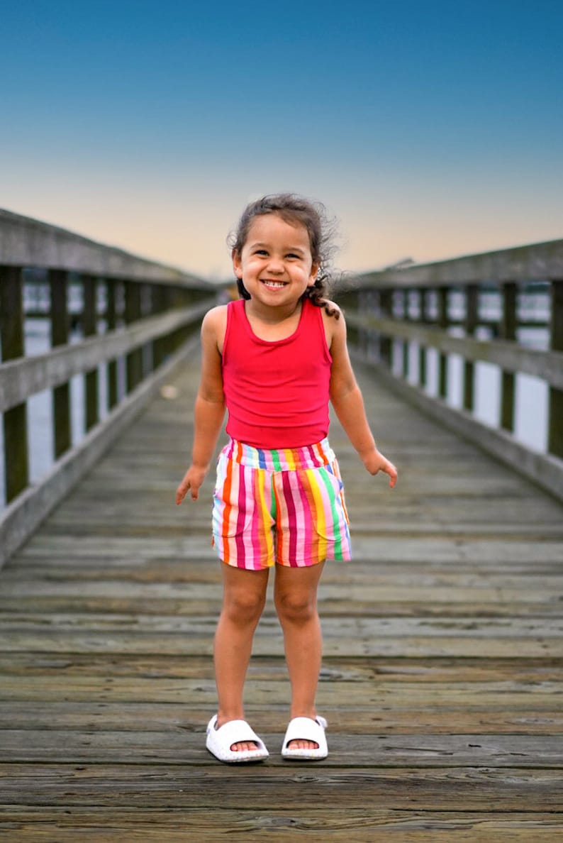 May include: A young girl wearing a red tank top and rainbow striped shorts smiles while standing on a wooden pier.