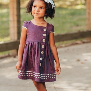 May include: A young girl in a plum-coloured dress with large buttons and white scalloped trim. The dress has short sleeves with white lace trim. She wears a white bow in her hair and stands barefoot outdoors.