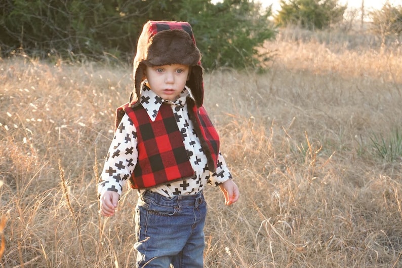 May include: A young child wearing a red and black plaid vest over a white shirt with black crosses, a brown fur-trimmed hat, and blue jeans. The child is standing in a field of tall grass.