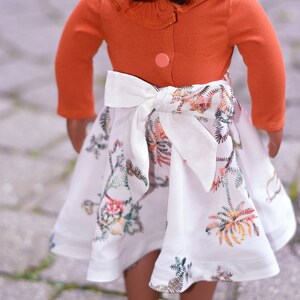 May include: A doll wearing a white dress with a floral pattern and a rust-colored long-sleeved shirt with a ruffled collar. The dress has a white bow tied at the waist.