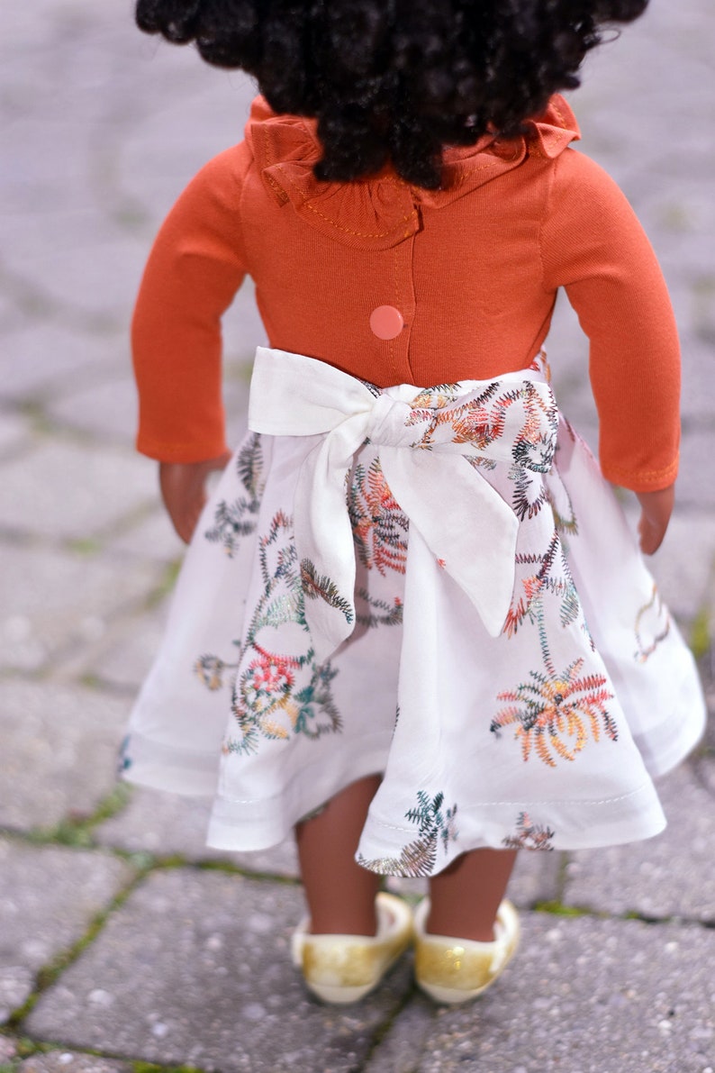 May include: A doll wearing a white dress with a floral pattern and a rust-colored long-sleeved shirt with a ruffled collar. The dress has a white bow tied at the waist.