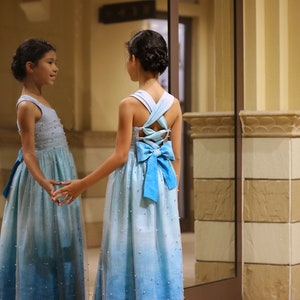 May include: A young girl in a light blue ombre dress with a blue bow on the back. The dress is covered in small white beads. She is standing in front of a mirror.