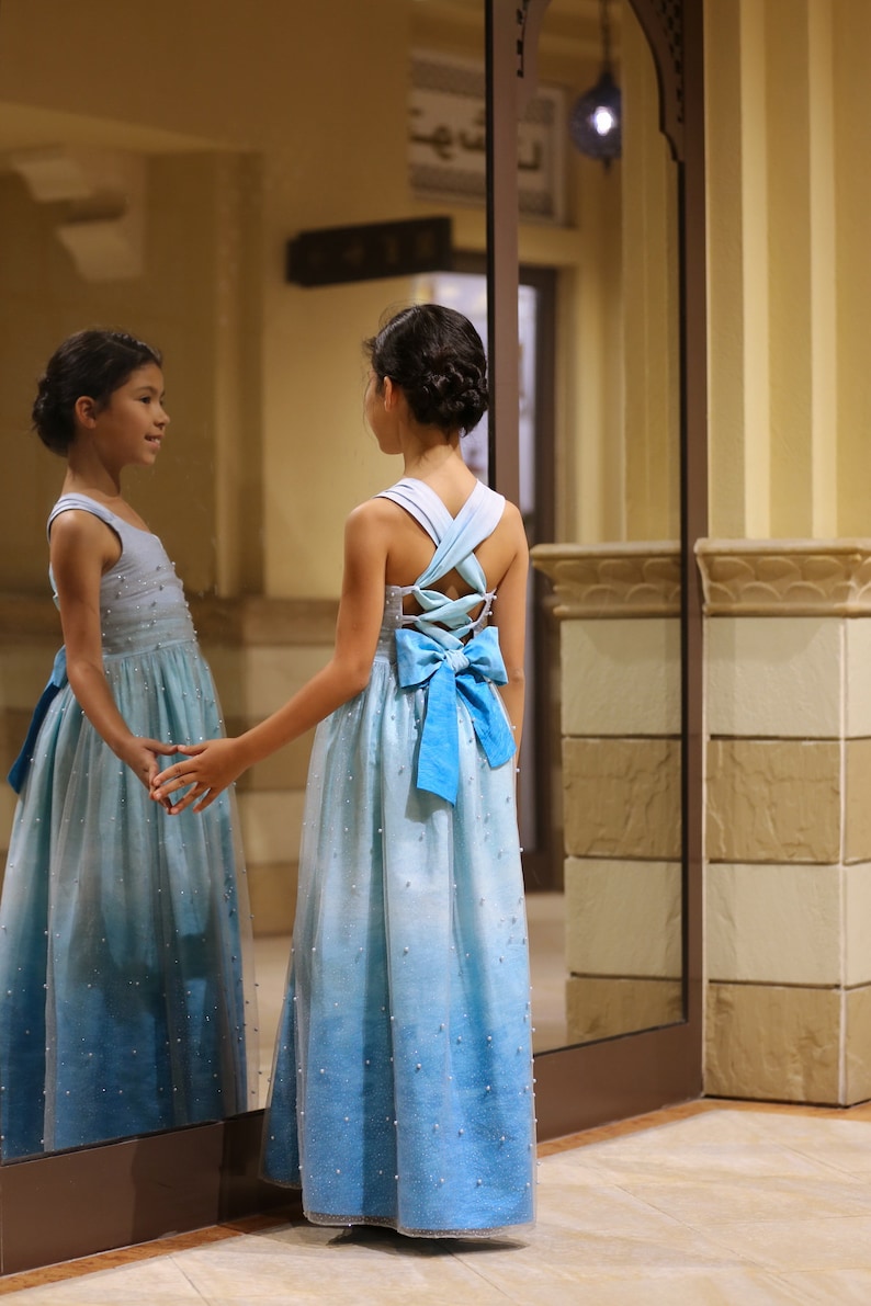 May include: A young girl in a light blue ombre dress with a blue bow on the back. The dress is covered in small white beads. She is standing in front of a mirror.