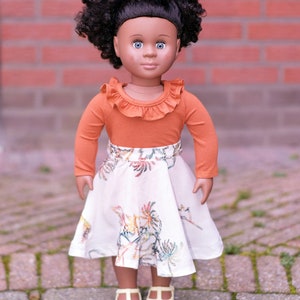 May include: A doll with brown skin and curly black hair is wearing a long-sleeved orange shirt with a ruffled neckline and a white skirt with a floral pattern. The doll is wearing gold shoes and is standing on a brick walkway.