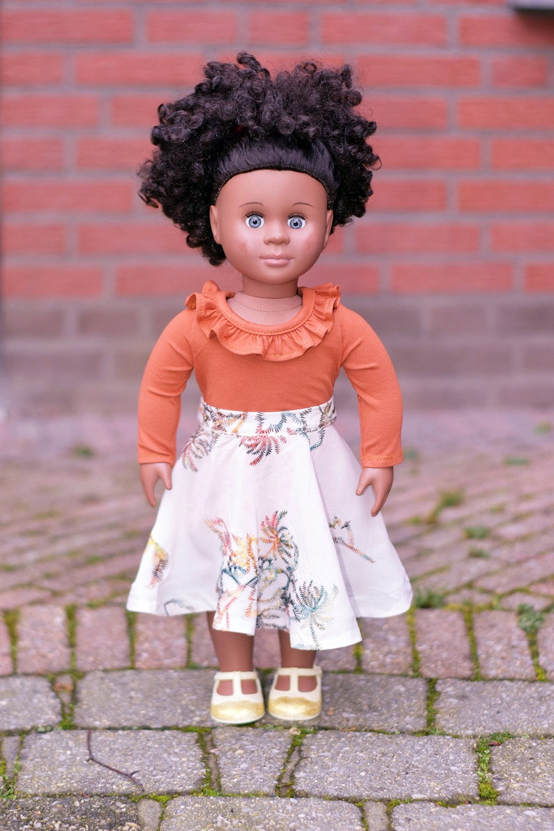 May include: A doll with brown skin and curly black hair is wearing a long-sleeved orange shirt with a ruffled neckline and a white skirt with a floral pattern. The doll is wearing gold shoes and is standing on a brick walkway.
