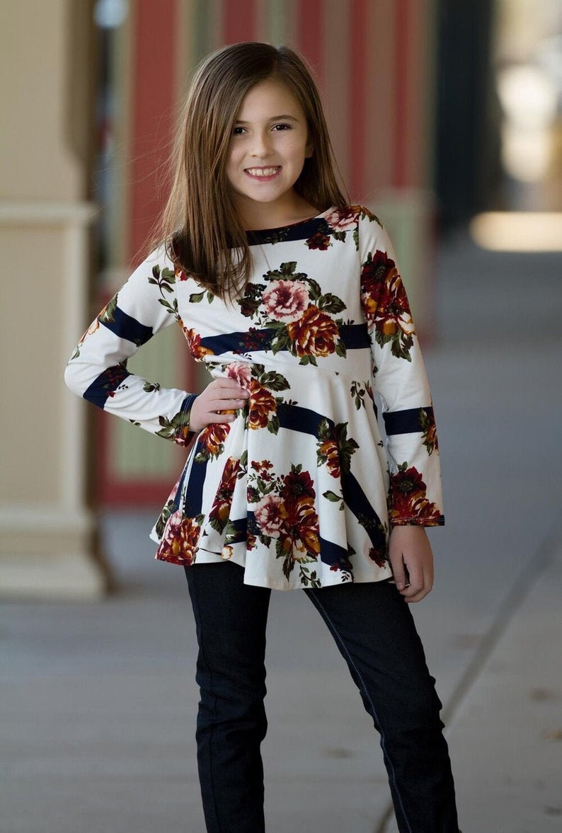 May include: A young person wearing a white and navy blue long-sleeved shirt with a floral print. The shirt has a ruffled hem and is paired with dark blue jeans.