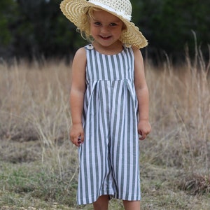 May include: A young girl wearing a striped jumpsuit with a straw hat and gold sandals. She is standing in a field of tall grass.