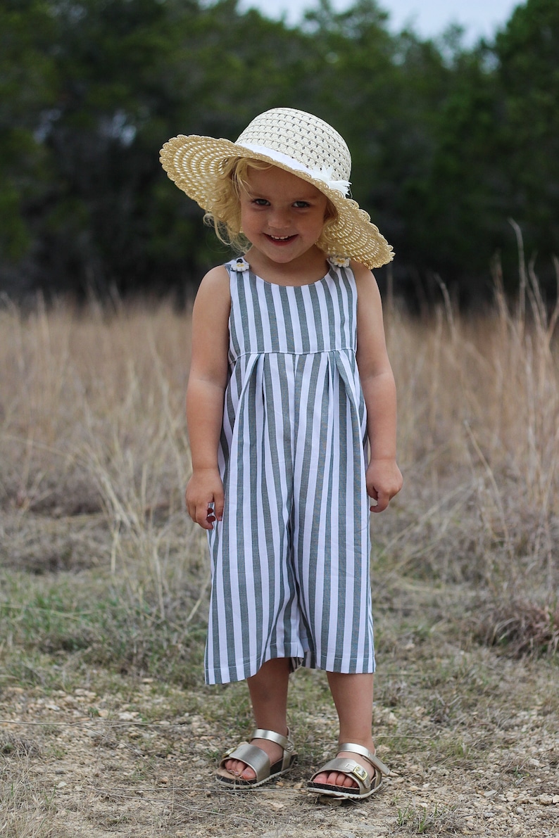 May include: A young girl wearing a striped jumpsuit with a straw hat and gold sandals. She is standing in a field of tall grass.