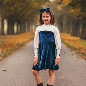 May include: A young person wearing a white and blue dress with a velvet skirt and a blue bow headband. They are standing on a paved path with trees on either side.
