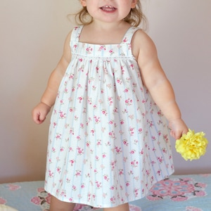 Pu&ograve; includere: Una bambina che indossa un vestito bianco con stampa floreale rosa e blu e sottili strisce blu. &Egrave; in piedi su un trapuntino a motivi blu e rosa e tiene un fiore giallo.