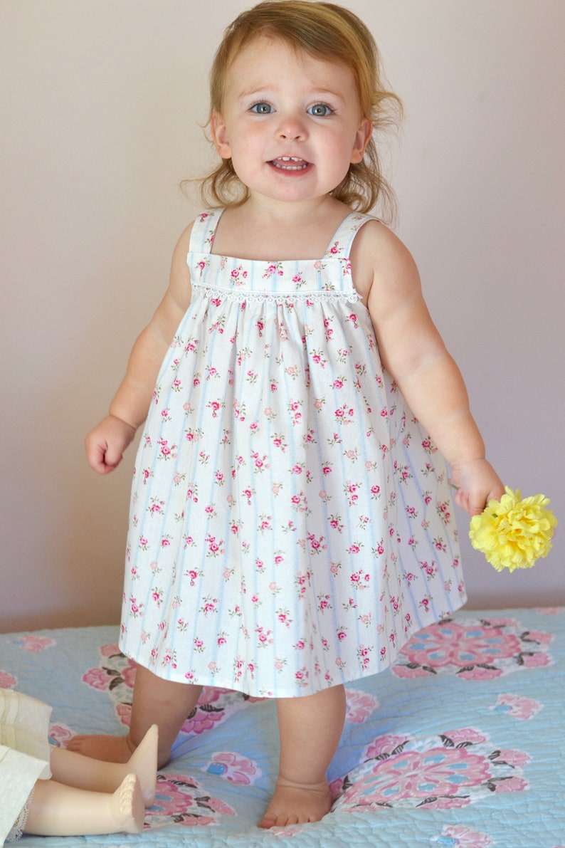 Pu&ograve; includere: Una bambina che indossa un vestito bianco con stampa floreale rosa e blu e sottili strisce blu. &Egrave; in piedi su un trapuntino a motivi blu e rosa e tiene un fiore giallo.