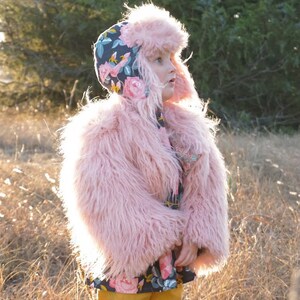 May include: A young child wearing a pink faux fur coat and a floral patterned hat. The child is standing in a field of tall grass.