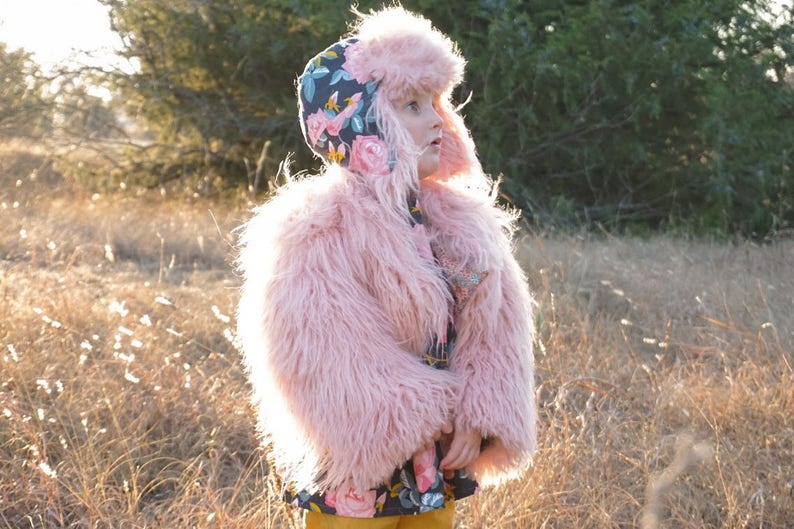 May include: A young child wearing a pink faux fur coat and a floral patterned hat. The child is standing in a field of tall grass.