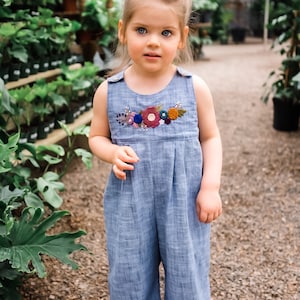 May include: A young girl wearing a light blue denim jumpsuit with embroidered flowers in shades of red, pink, blue, and green. The jumpsuit has a sleeveless design and a round neckline.