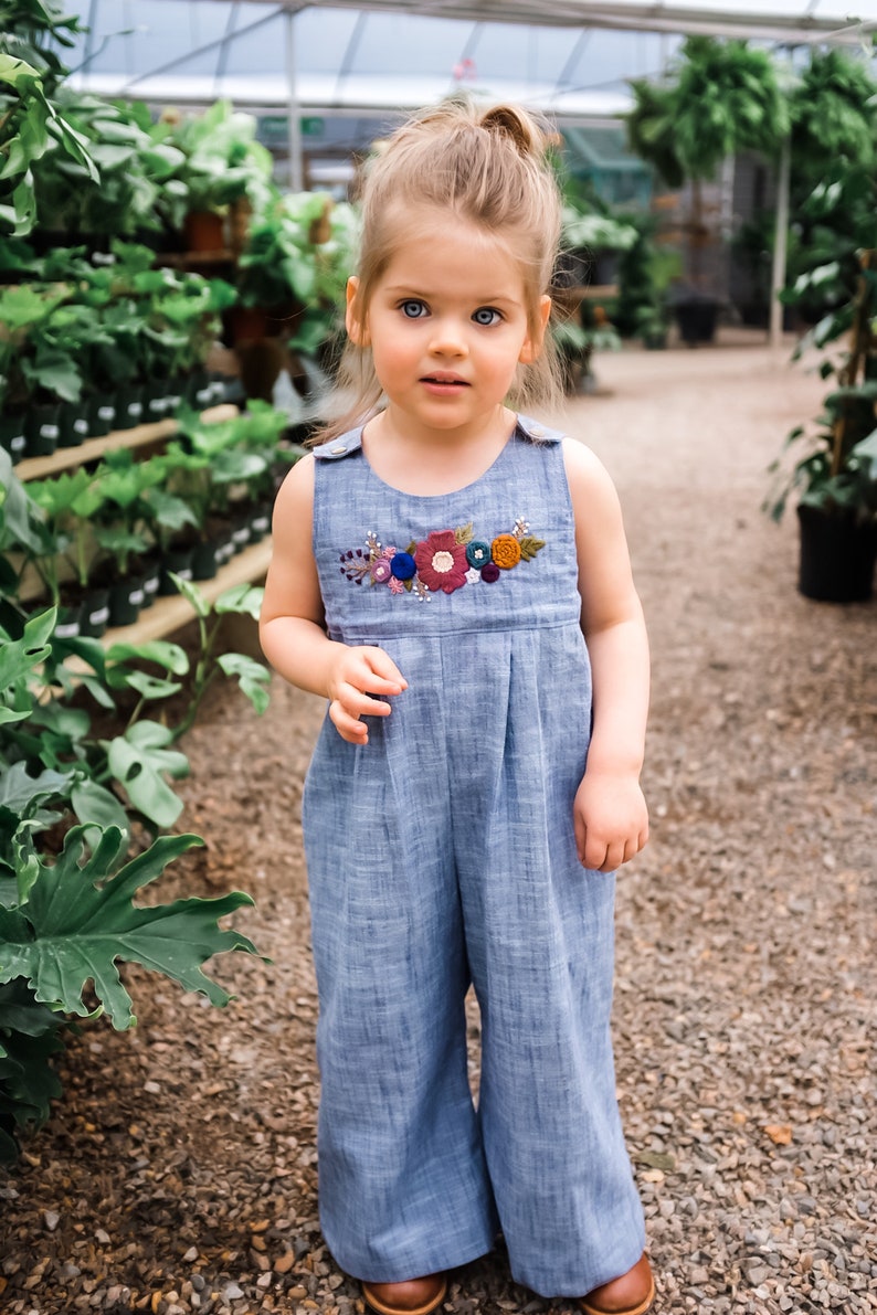May include: A young girl wearing a light blue denim jumpsuit with embroidered flowers in shades of red, pink, blue, and green. The jumpsuit has a sleeveless design and a round neckline.