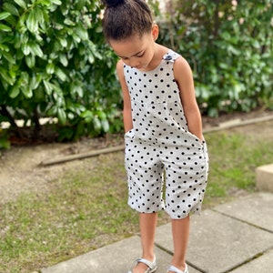May include: A young girl wearing a white and black polka dot jumpsuit with a single button on the strap. She is standing on a gray sidewalk with green grass in the background.
