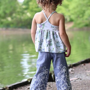 May include: A young girl wearing a white floral print tank top and blue denim pants with white embroidered flowers. The pants have a wide leg style.