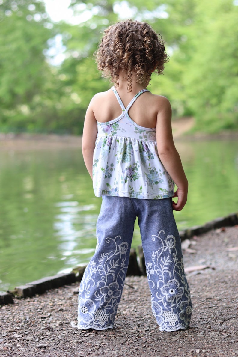 May include: A young girl wearing a white floral print tank top and blue denim pants with white embroidered flowers. The pants have a wide leg style.