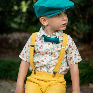 May include: A young boy wearing a teal newsboy cap, a white button-down shirt with a floral pattern, a teal bow tie, yellow suspenders, and yellow shorts. He is sitting on a tree trunk.