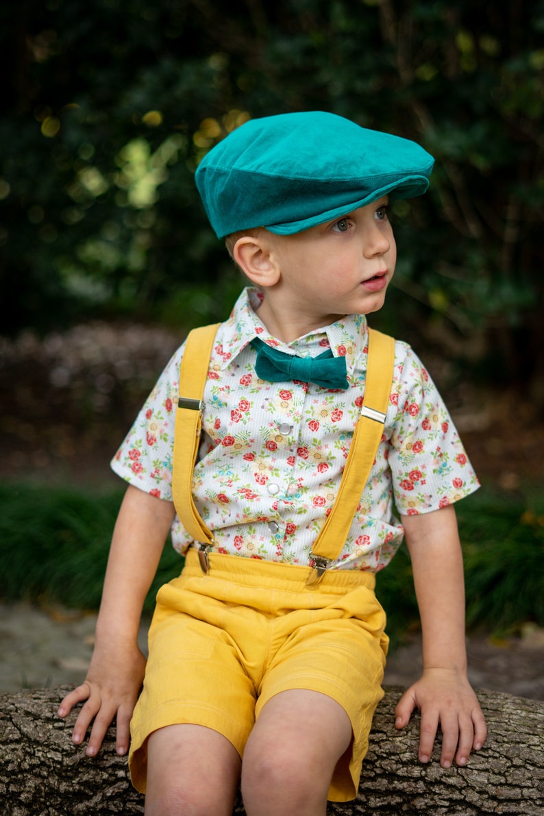 May include: A young boy wearing a teal newsboy cap, a white button-down shirt with a floral pattern, a teal bow tie, yellow suspenders, and yellow shorts. He is sitting on a tree trunk.