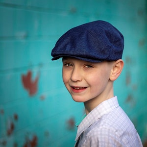 May include: A young boy wearing a blue newsboy cap and a white and blue plaid shirt smiles at the camera. He is standing in front of a turquoise wall.