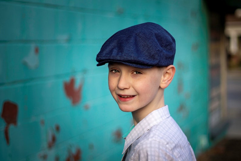 May include: A young boy wearing a blue newsboy cap and a white and blue plaid shirt smiles at the camera. He is standing in front of a turquoise wall.