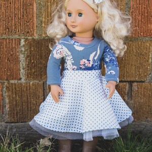 May include: A doll wearing a blue and white polka dot dress with a floral print top and a white bow in her hair. The doll is standing in front of a brick wall.