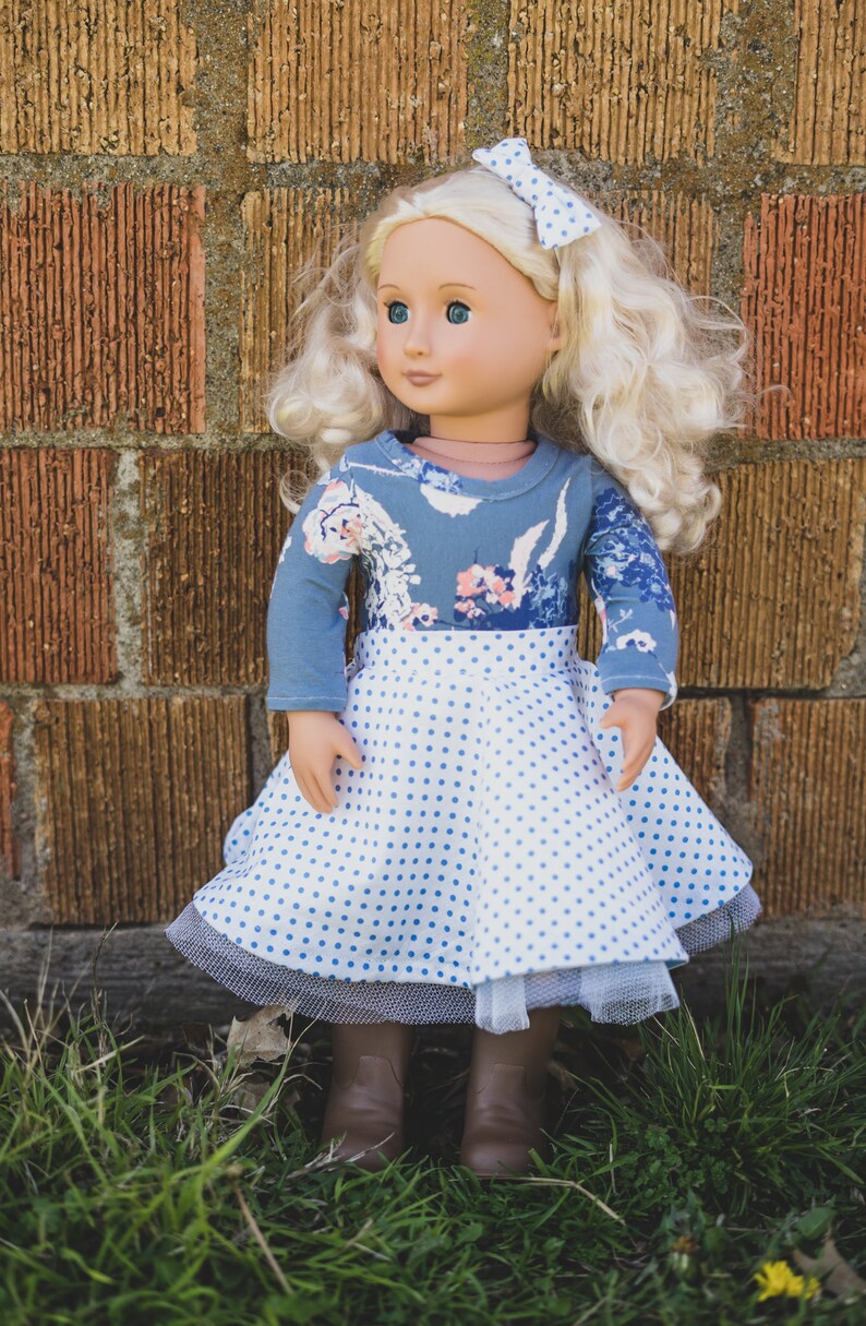 May include: A doll wearing a blue and white polka dot dress with a floral print top and a white bow in her hair. The doll is standing in front of a brick wall.