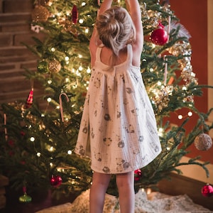 Pu&ograve; includere: Una bambina con un vestito bianco con un motivo di piccoli animali decora un albero di Natale con un ornamento "NOEL" rosso e nero. L'albero &egrave; decorato con luci, ornamenti e bastoncini di zucchero.