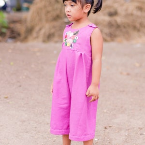 May include: A young girl wearing a pink sleeveless jumpsuit with a floral embroidered design. She is standing on a dirt path with a pile of hay in the background.