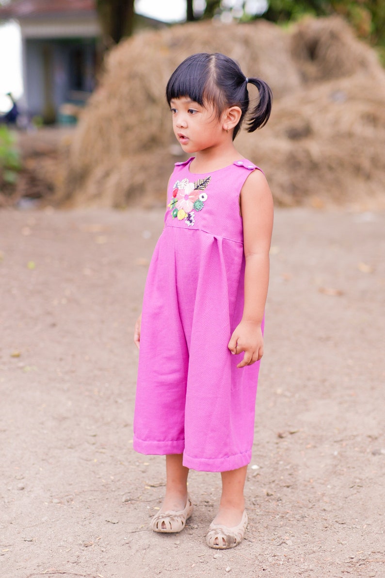 May include: A young girl wearing a pink sleeveless jumpsuit with a floral embroidered design. She is standing on a dirt path with a pile of hay in the background.