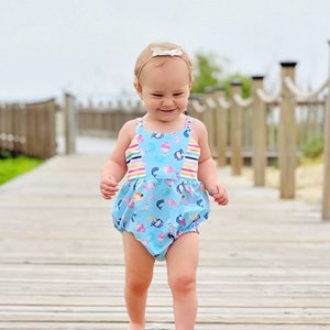 May include: A baby girl wearing a light blue romper with a colorful pattern of sea creatures and ice cream cones. The romper has striped accents and a ruffled bottom. She is walking on a wooden boardwalk, wearing a headband with a bow.