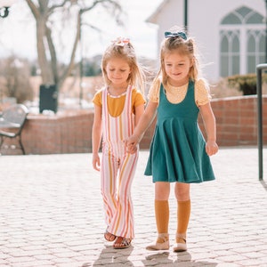 May include: Two young girls wearing colorful clothing walk hand-in-hand on a brick pathway. One girl wears a teal dress and yellow socks, while the other wears a pink and yellow striped jumpsuit.