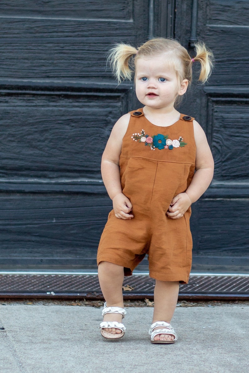 May include: A young child wearing a brown overall with embroidered flowers. The overall has a button closure at the neck. The child is wearing white sandals and has their hair in pigtails.