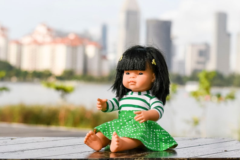 May include: A green and white striped shirt with a green polka dot skirt. The doll has black hair and brown eyes. The doll is sitting on a wooden table.