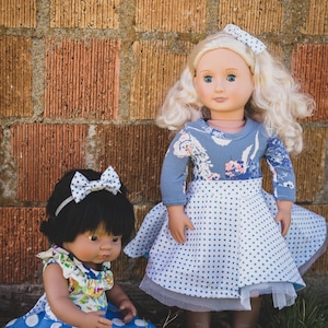 May include: Two dolls wearing blue and white polka dot dresses. The doll on the left has black hair and is wearing a floral print top. The doll on the right has blonde hair and is wearing a blue floral print top. Both dolls are standing in front of a brick wall.