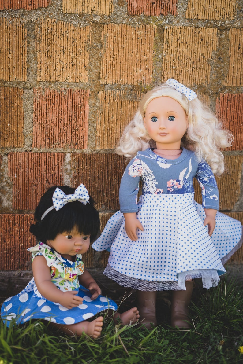 May include: Two dolls wearing blue and white polka dot dresses. The doll on the left has black hair and is wearing a floral print top. The doll on the right has blonde hair and is wearing a blue floral print top. Both dolls are standing in front of a brick wall.